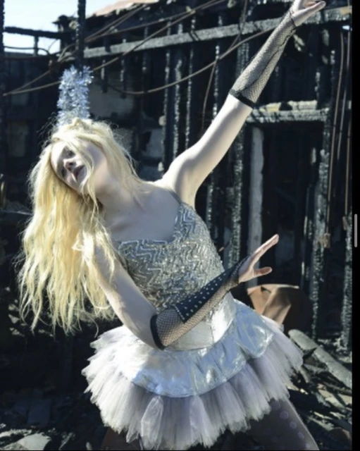 Blonde woman in a shimmering dress and fishnet gloves striking a pose in front of a burned-out building, artistic fashion shoot.