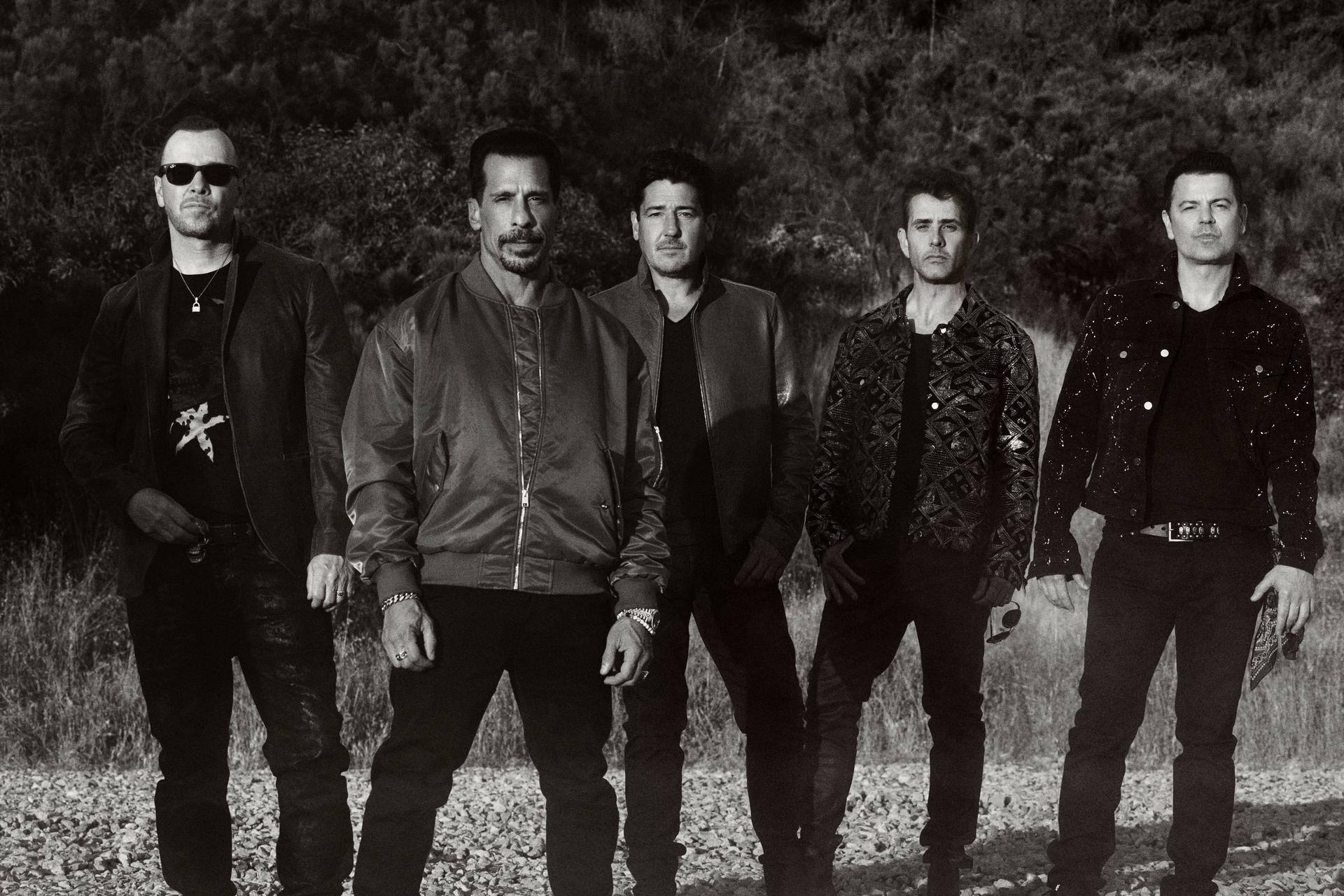 Five men in jackets pose outdoors in a black-and-white shot. One man wears sunglasses, another a patterned jacket. The group is in a field.