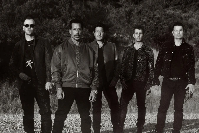 Five men in jackets pose outdoors in a black-and-white shot. One man wears sunglasses, another a patterned jacket. The group is in a field.