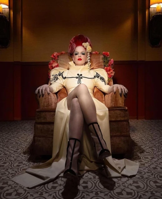 Woman in a yellow dress and red wig sitting in a chair with floral accents and patterned floors, creating a vintage, theatrical aesthetic.