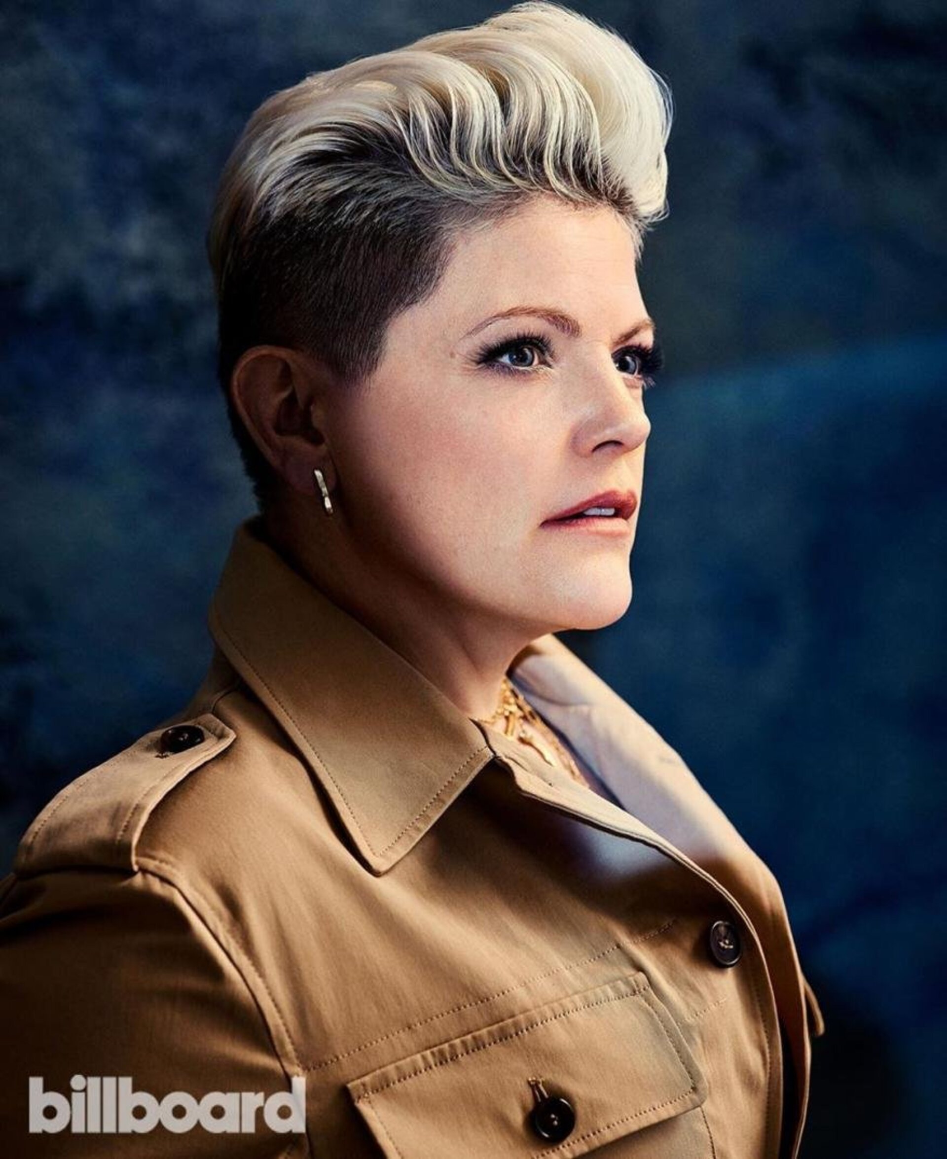 Headshot of a blonde woman with short hair wearing a tan trench coat. She is positioned in front of a dark, abstract background.
