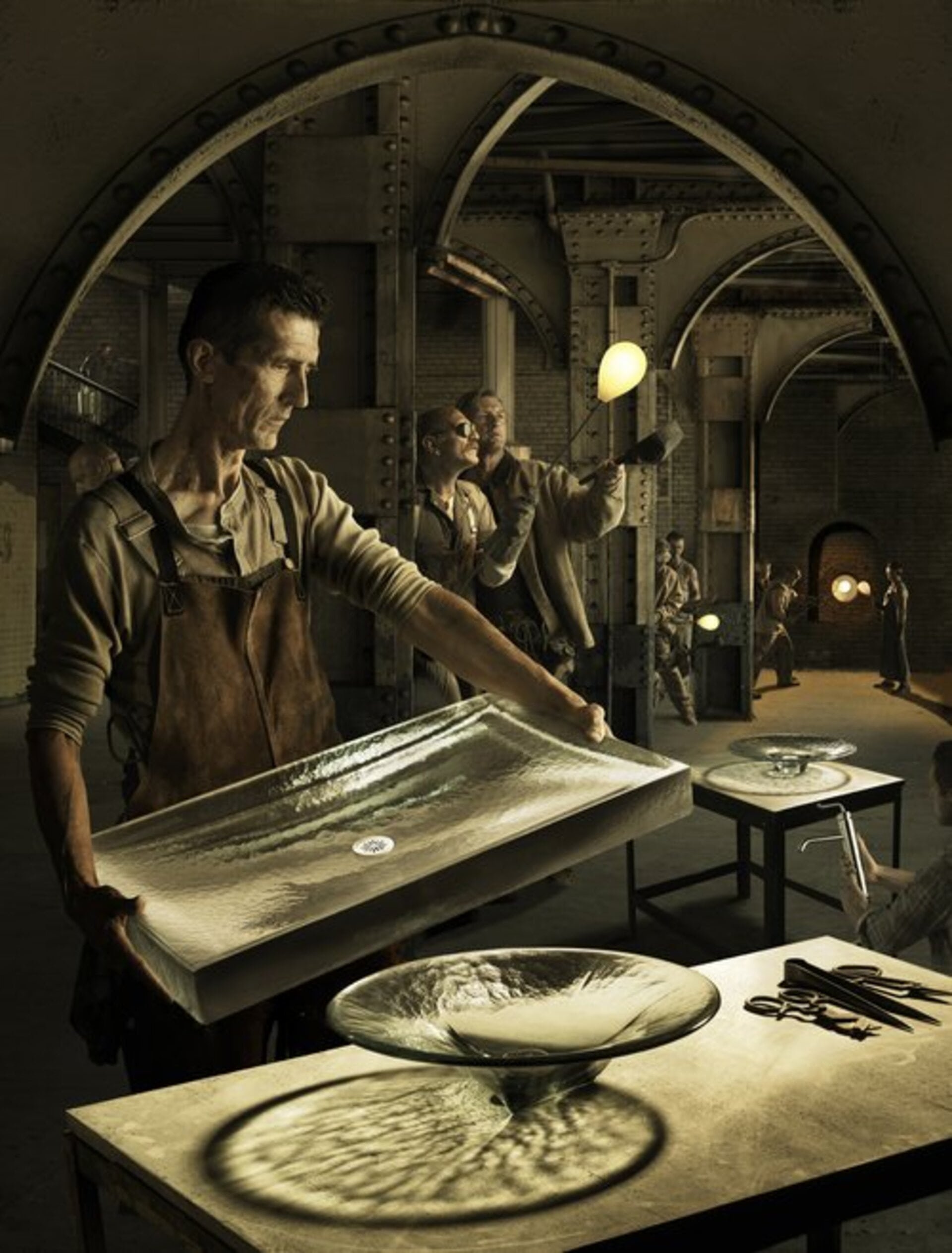 Craftsman holding a textured glass basin in an industrial glassblowing factory, with a group of people working with molten glass.