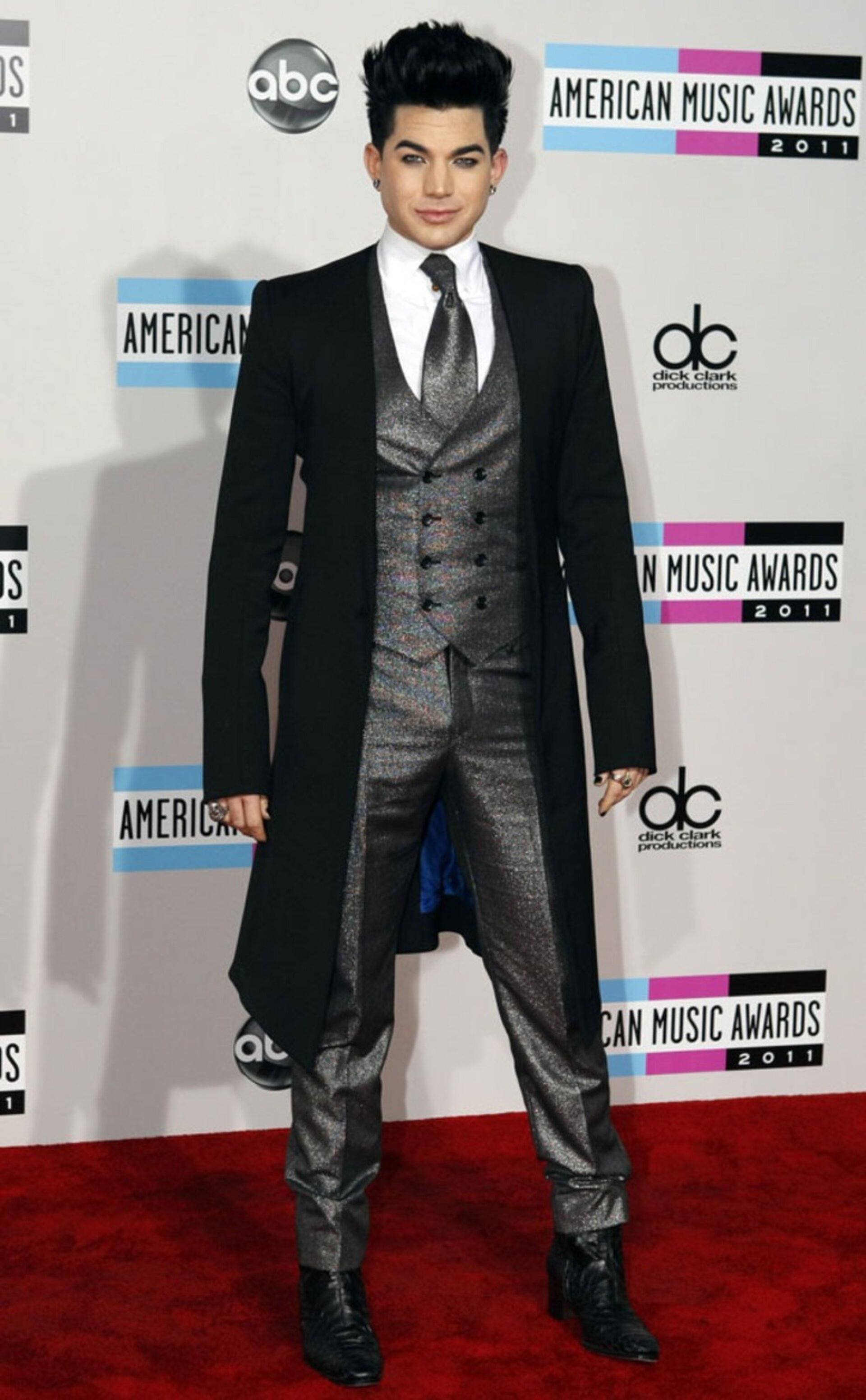Adam Lambert poses on a red carpet at the American Music Awards in a black tailcoat, grey vest, and patterned pants, smiling slightly.