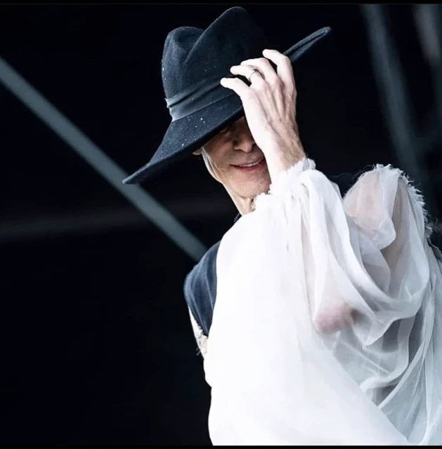 A person wearing a wide-brimmed hat and white, flowing blouse smiles with a hand near the brim of the hat against a black background.
