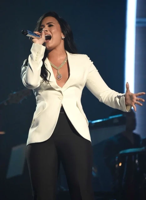 A celebrity singer performing on stage wearing a white blazer, black pants, and a large statement necklace with a microphone in hand.