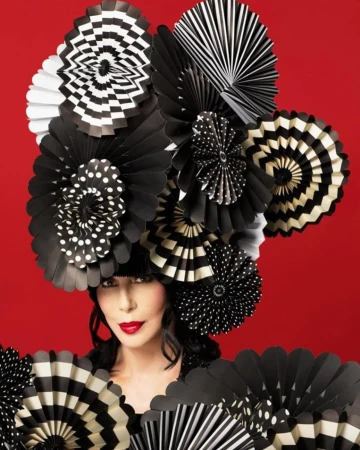 Woman in artistic headdress of black-and-white patterned paper fans against a red background. She wears red lipstick and dark clothing.