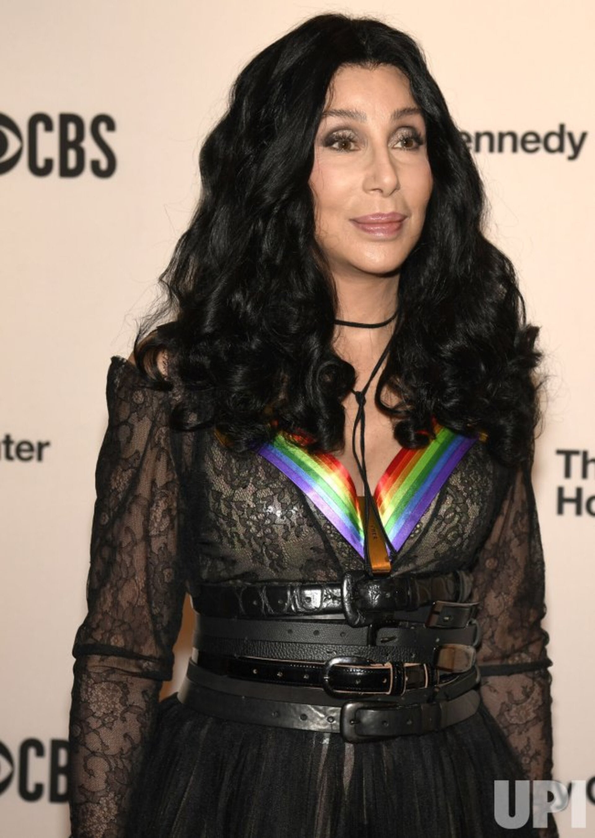 Close-up of Cher with long black curly hair, wearing a rainbow-colored pendant, and a black lace dress, at an indoor event.