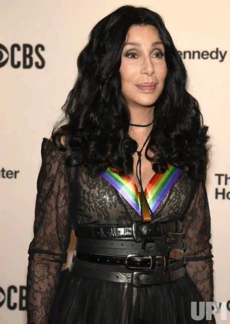 Close-up of Cher with long black curly hair, wearing a rainbow-colored pendant, and a black lace dress, at an indoor event. 