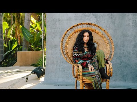 Cher sits in a wicker peacock chair in front of a gray backdrop with a plant and lighting equipment visible in the background.