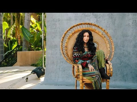 Cher sits in a wicker peacock chair in front of a gray backdrop with a plant and lighting equipment visible in the background.