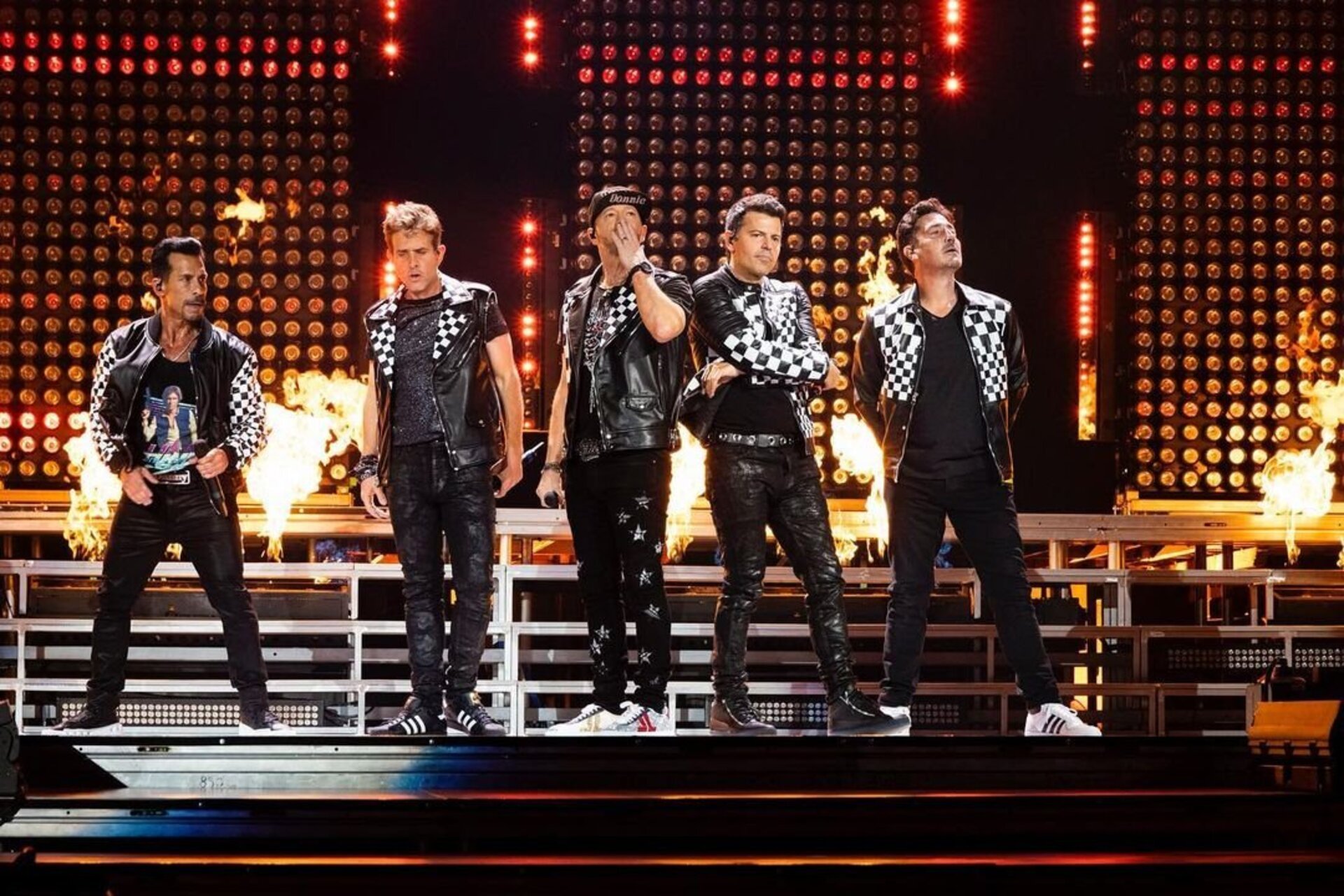 Five men in black leather jackets with checkerboard accents perform on a stage with a fiery backdrop, illuminated by stage lighting.