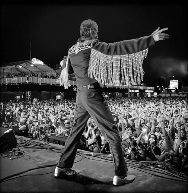 Black and white concert photograph features a male musician on stage in a fringed jacket, performing for a large crowd at an outdoor event.