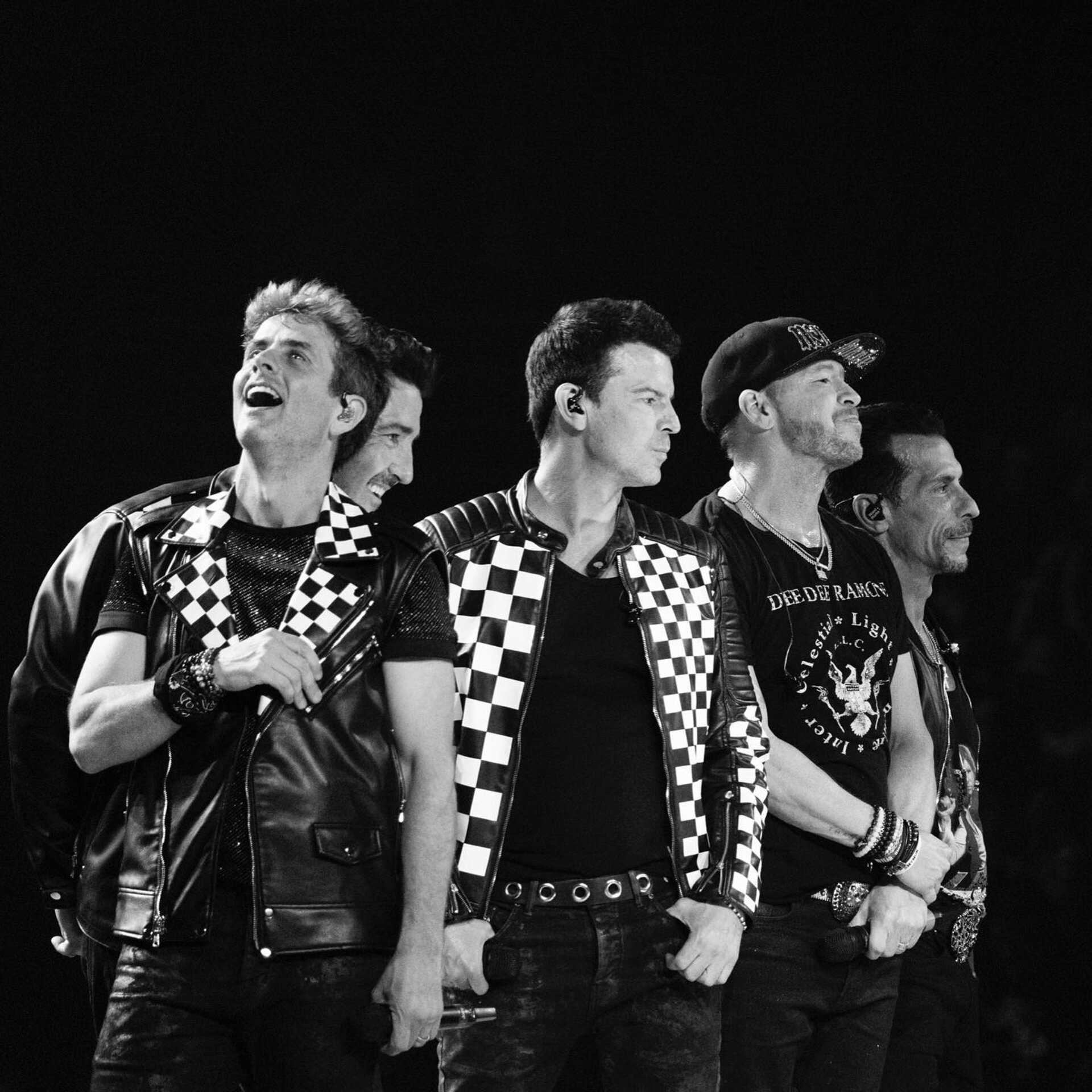 Monochrome shot of New Kids on the Block performing onstage, wearing black leather jackets and pants, smiling with stage lighting.