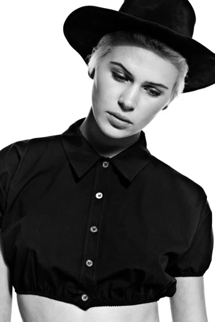 Black and white portrait of a young woman with platinum blonde hair wearing a black hat and a short black blouse, in a photo studio.