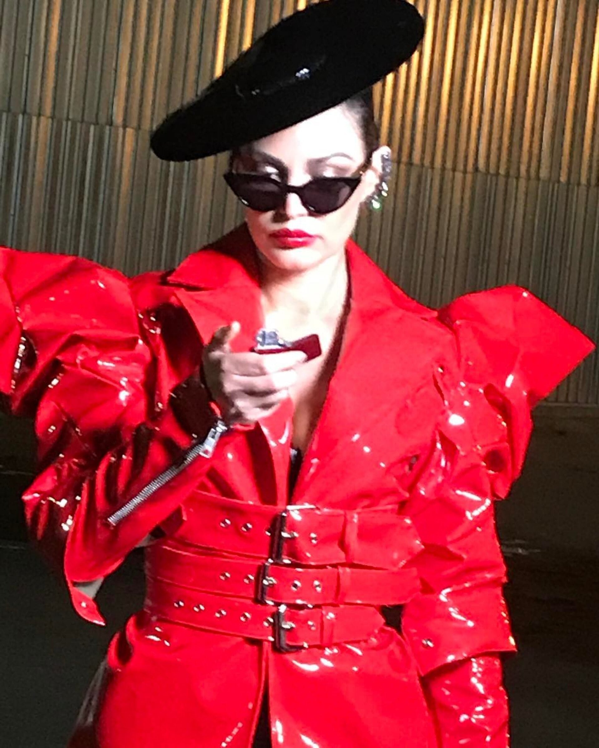 Adult female in a shiny red coat and black hat holds a lighter in an indoor environment with minimalist background details. High fashion portrait.
