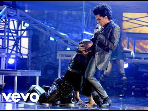 Adam Lambert performs on stage with two backup dancers in a shimmering suit under blue stage lights with an industrial metal set design.