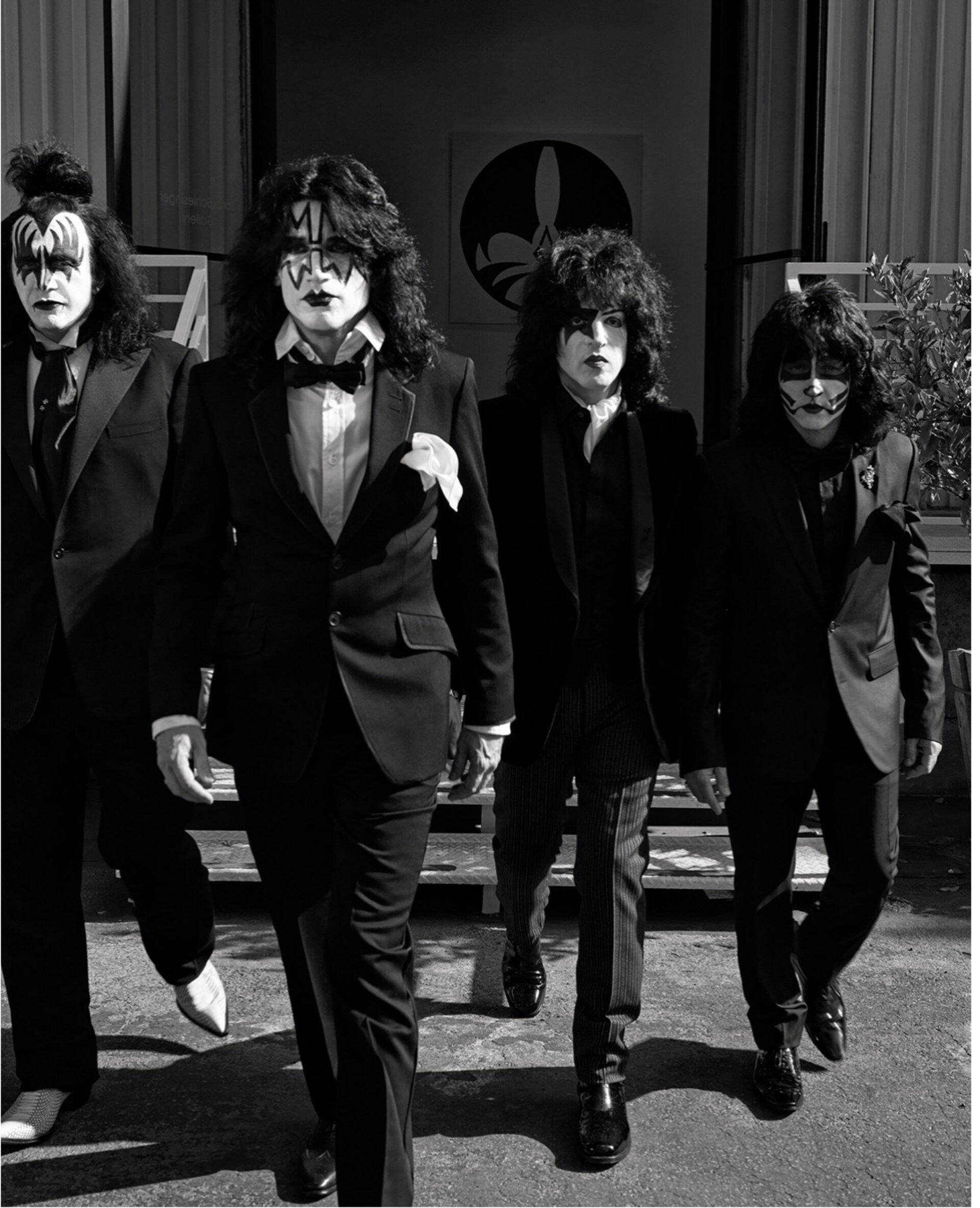 Four adult males dressed in black suits and KISS-style face makeup walk toward the camera in front of a building exterior with foliage.