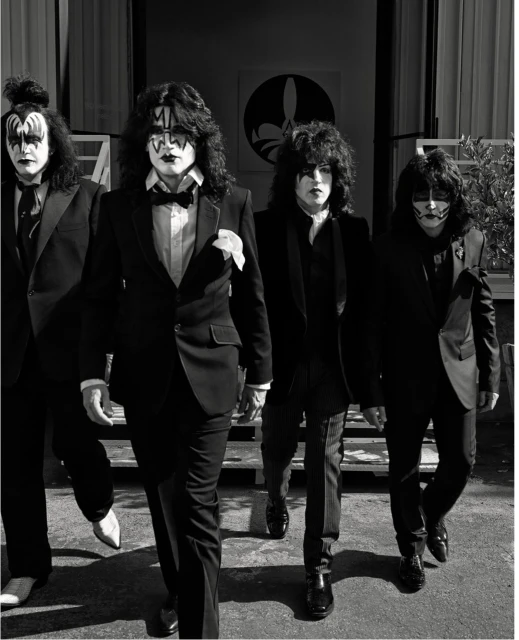Four adult males dressed in black suits and KISS-style face makeup walk toward the camera in front of a building exterior with foliage.