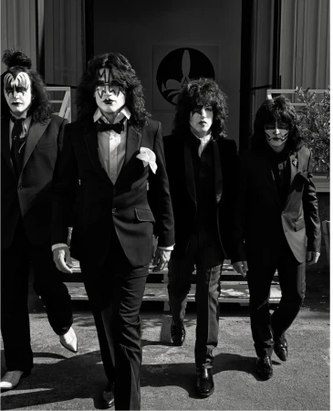 Four adult males dressed in black suits and KISS-style face makeup walk toward the camera in front of a building exterior with foliage.