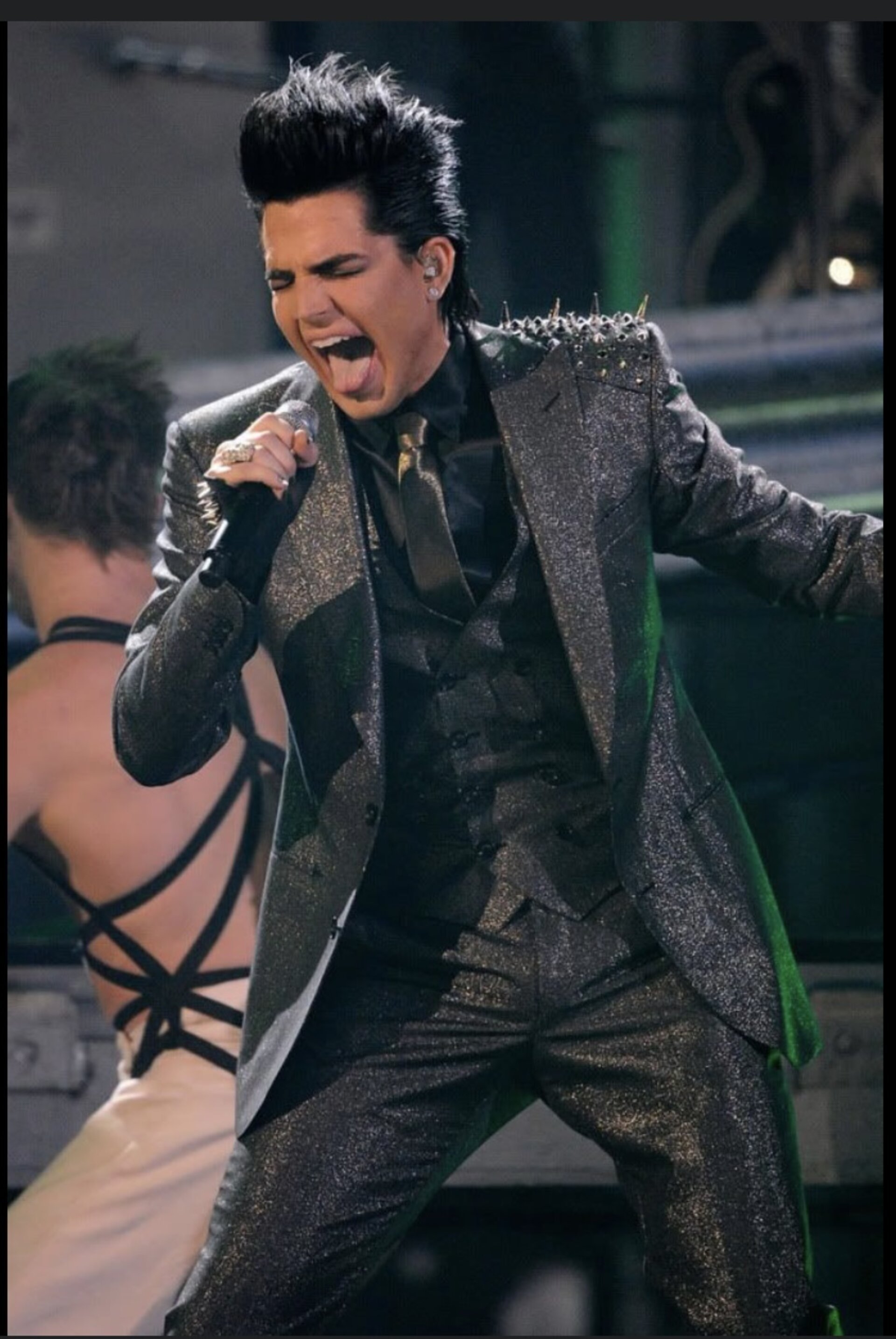 Adam Lambert, singer, performs in a glittery dark grey suit with spiked shoulder pads, singing with an open mouth, holding a microphone.