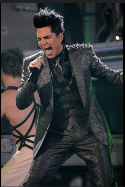 Adam Lambert, singer, performs in a glittery dark grey suit with spiked shoulder pads, singing with an open mouth, holding a microphone.