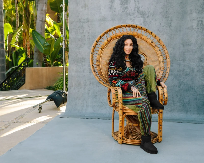 Cher sits in a wicker peacock chair outdoors, wearing a patterned sweater, green pants, and black boots, smiling against a grey backdrop.