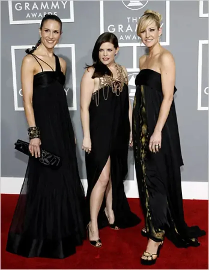 Three women in formal black gowns pose on a red carpet, attending the GRAMMY Awards, against a grey backdrop with the GRAMMY Awards logo.