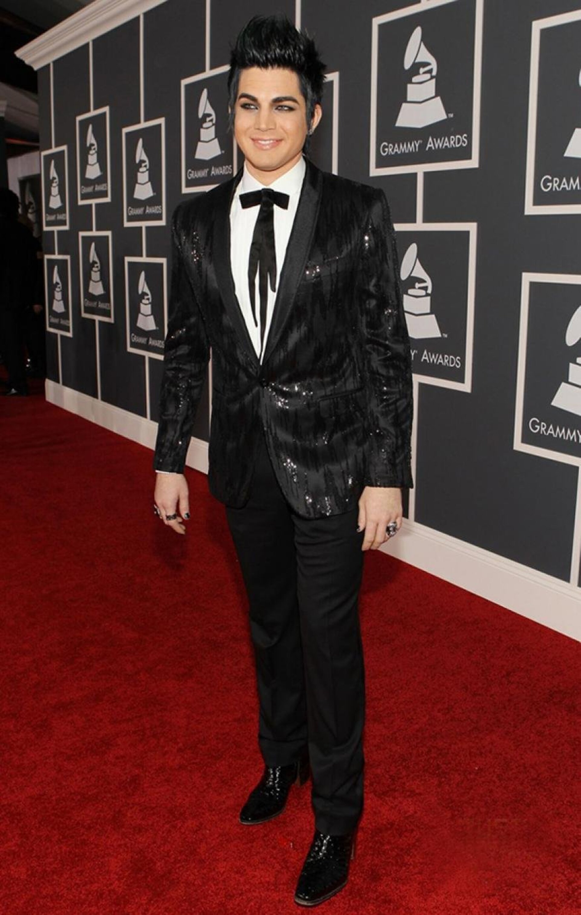 Adam Lambert poses on a red carpet wearing a black sequined suit with a bow tie and black shoes in front of a grey background.