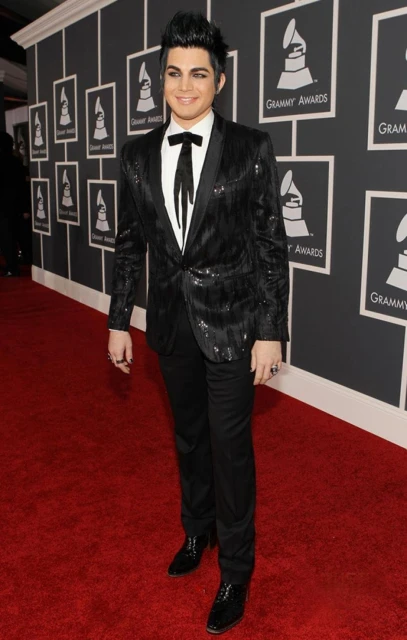 Adam Lambert poses on a red carpet wearing a black sequined suit with a bow tie and black shoes in front of a grey background.