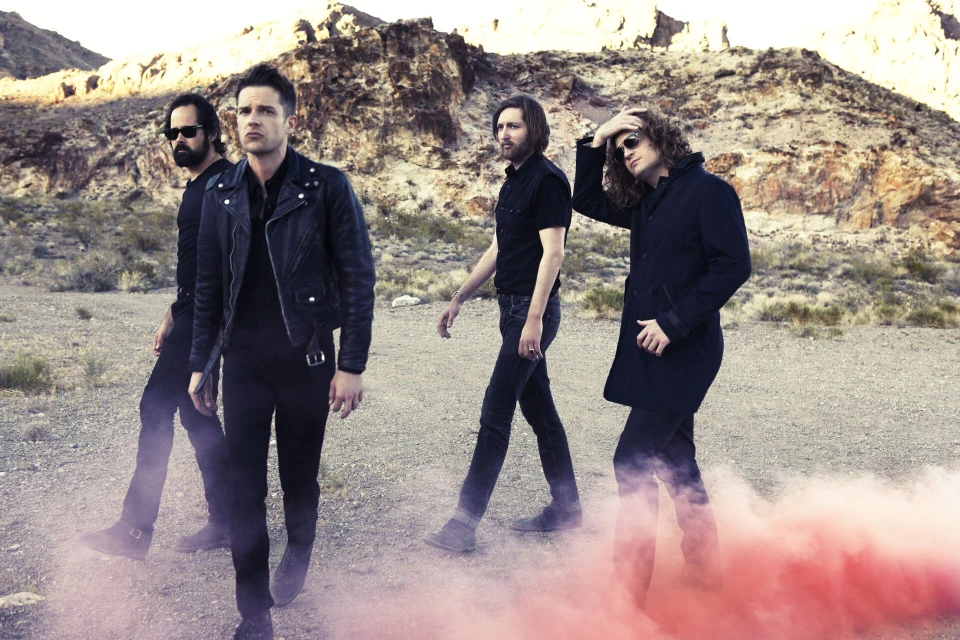 Four adult males walk through desert with red smoke, wearing dark clothing with mountains and sky in the background in a fashion editorial.