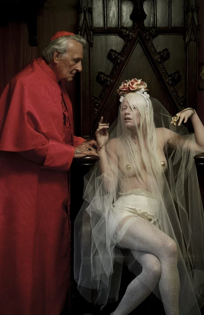 Striking image of an older male in red cardinal attire standing next to a woman in lingerie seated on a throne-like chair.
