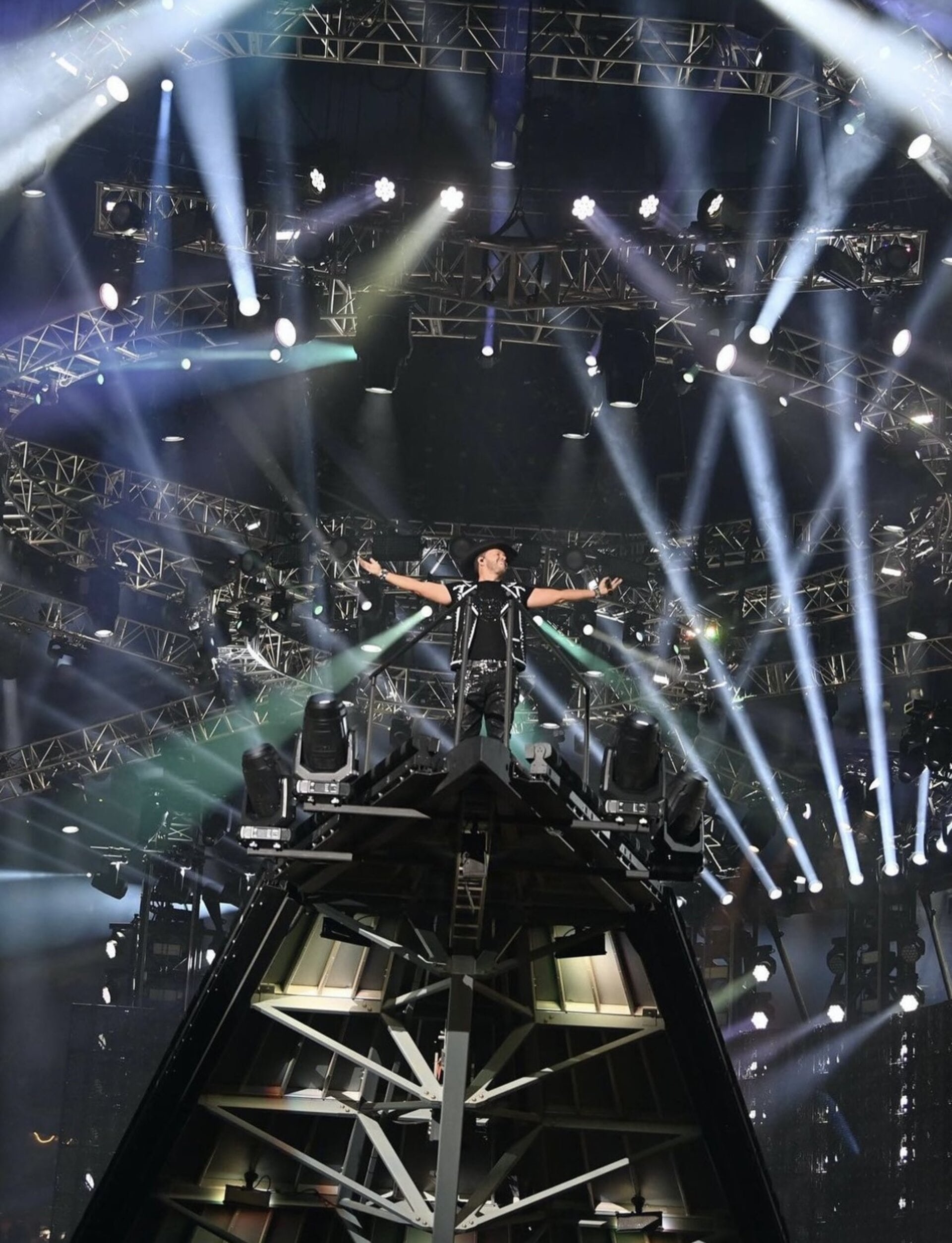 An adult male stands on a stage platform with his arms outstretched, bathed in bright spotlights and stage lighting, during a concert.