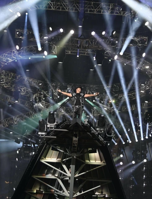 An adult male stands on a stage platform with his arms outstretched, bathed in bright spotlights and stage lighting, during a concert.