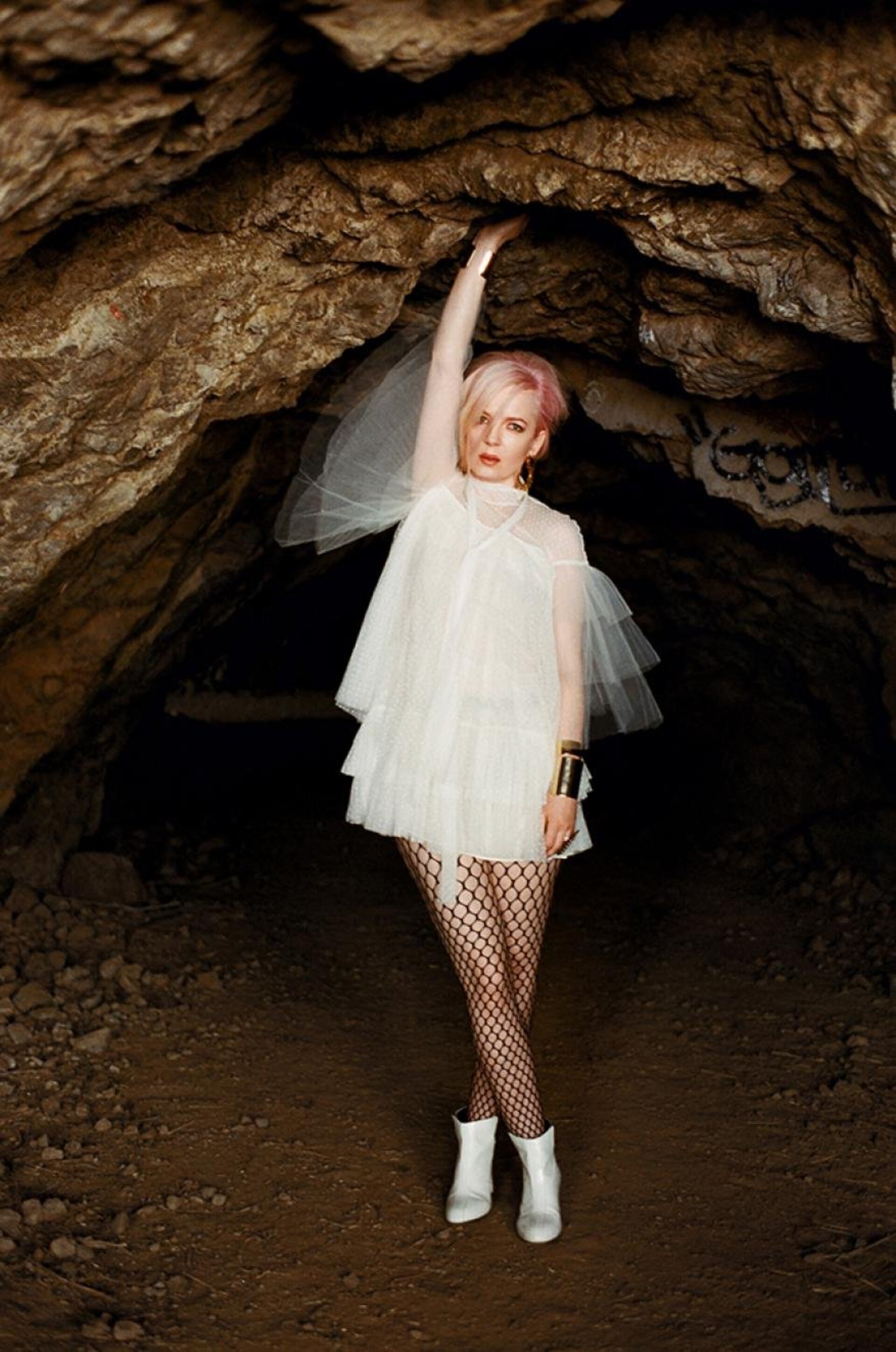 Woman in a white tulle dress and fishnet stockings poses inside a dark cave, holding onto the cave ceiling with her hand.
