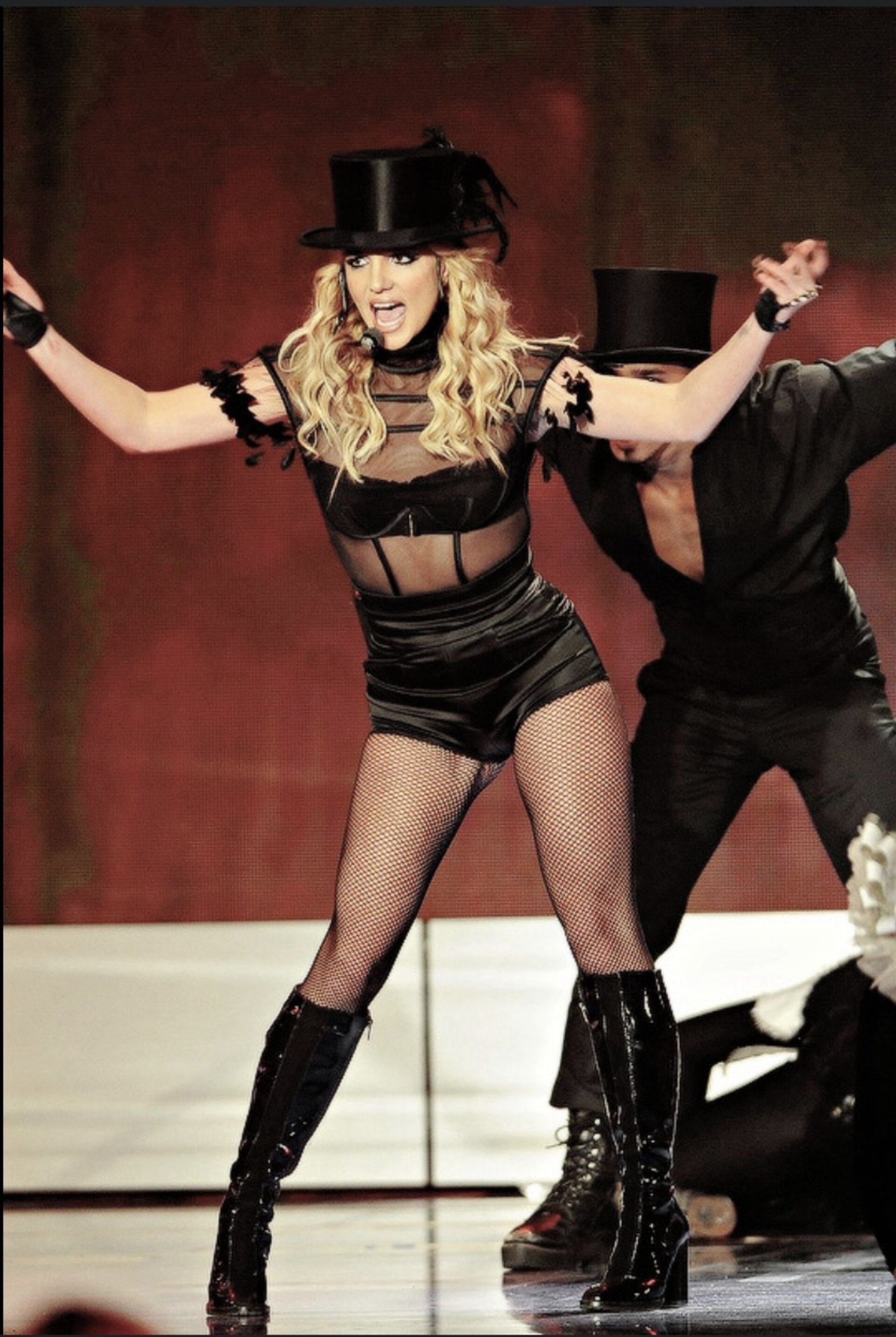 A performer in a black top hat, a sheer black top, black shorts, and patent leather boots is captured mid-performance on a red-lit stage.
