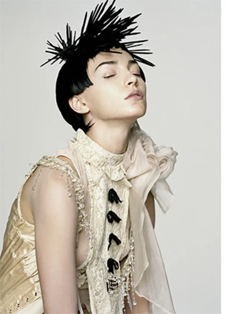Woman with spiky hair poses in a cream-colored lace outfit against a plain white background in a studio lighting environment.