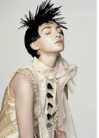 Woman with spiky hair poses in a cream-colored lace outfit against a plain white background in a studio lighting environment.