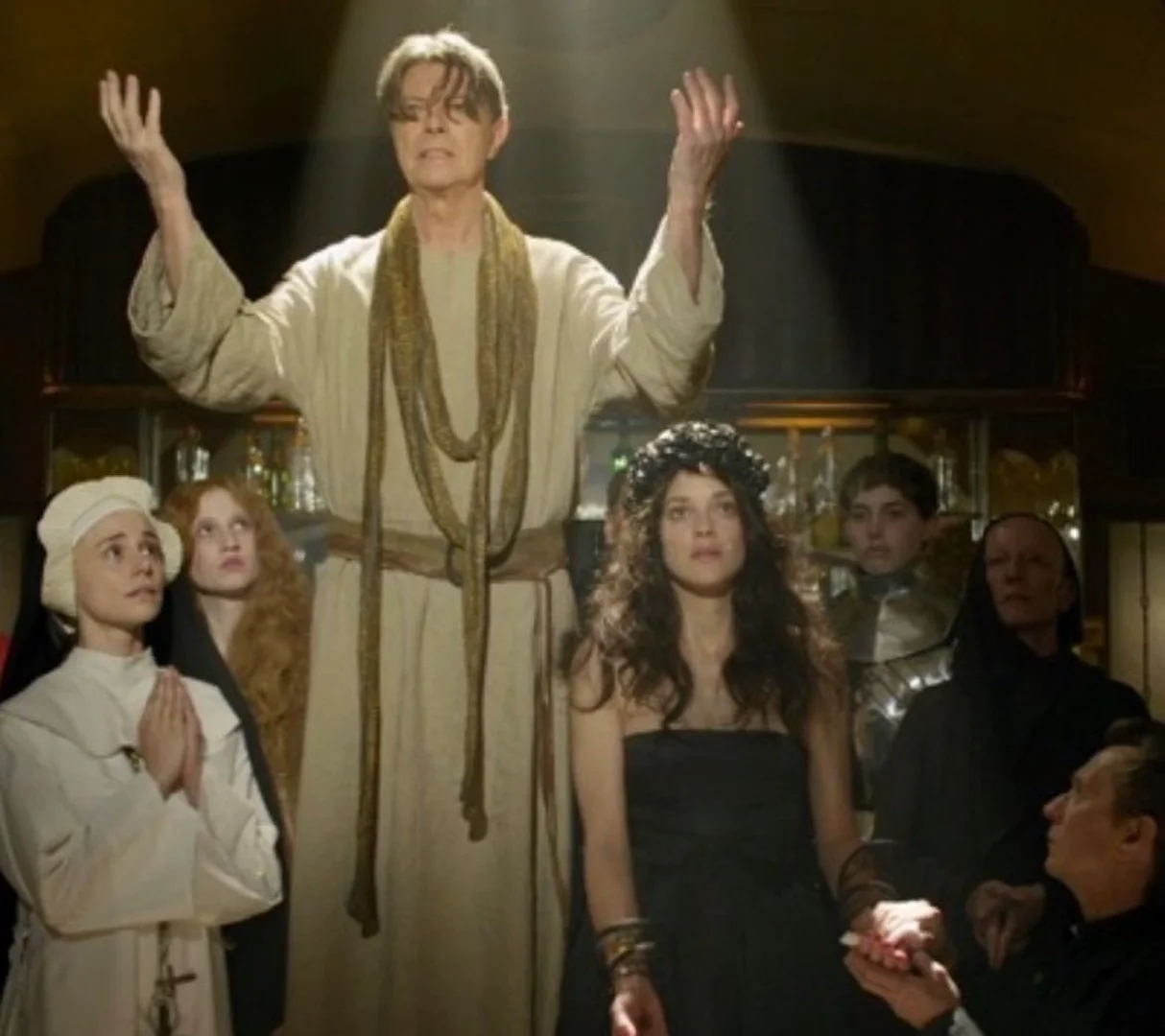 David Bowie stands with raised arms among a group of people in an ornate, dimly lit indoor setting with architectural details.