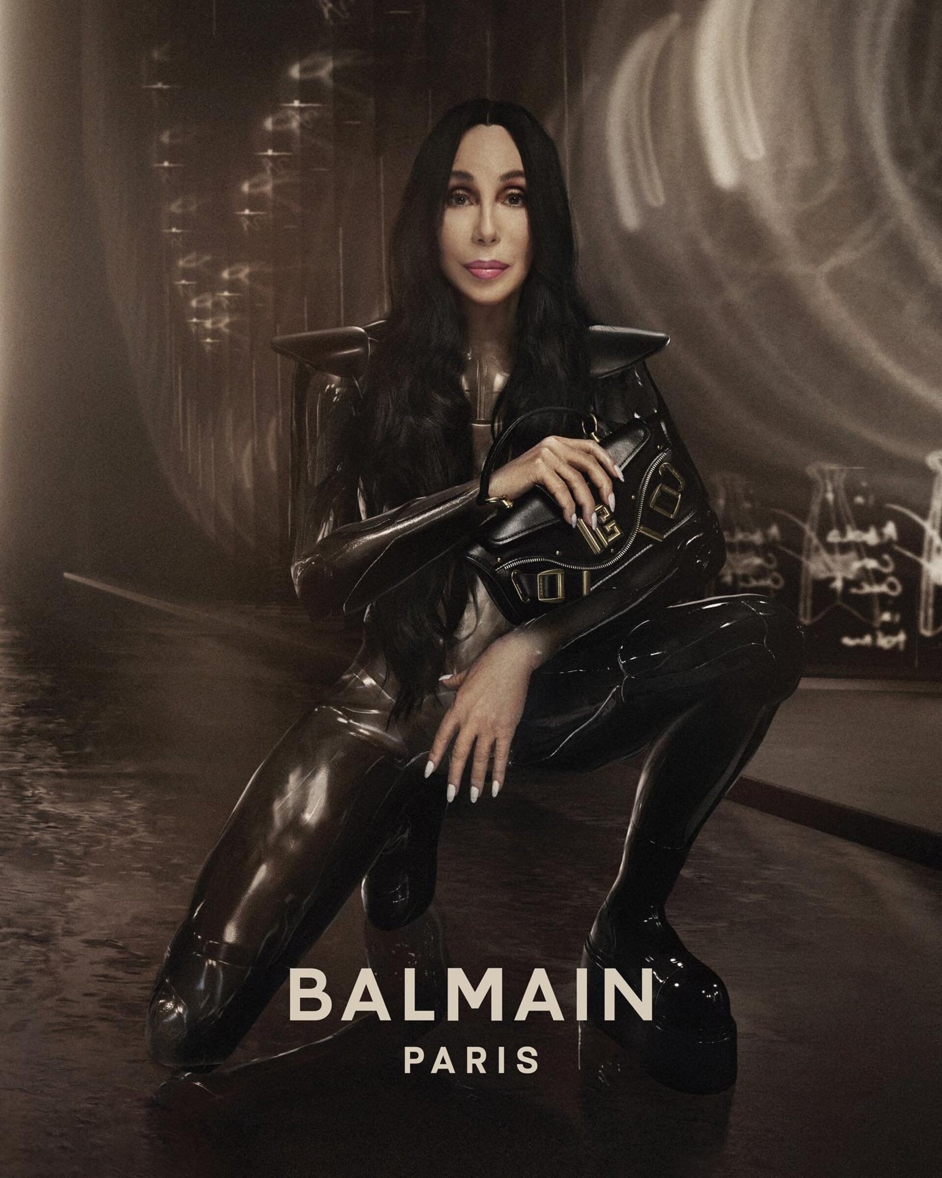 Singer Cher poses in glossy black outfit with Balmain handbag, crouching indoors with diffused light, for a fashion advertisement.