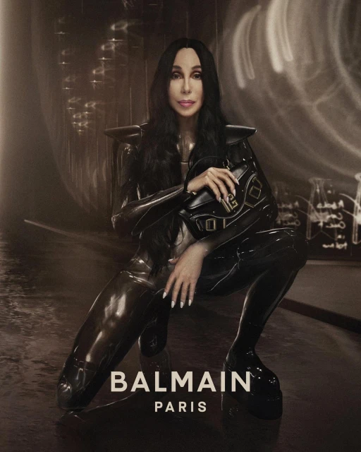 Singer Cher poses in glossy black outfit with Balmain handbag, crouching indoors with diffused light, for a fashion advertisement.