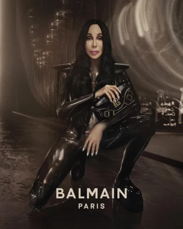 Singer Cher poses in glossy black outfit with Balmain handbag, crouching indoors with diffused light, for a fashion advertisement.