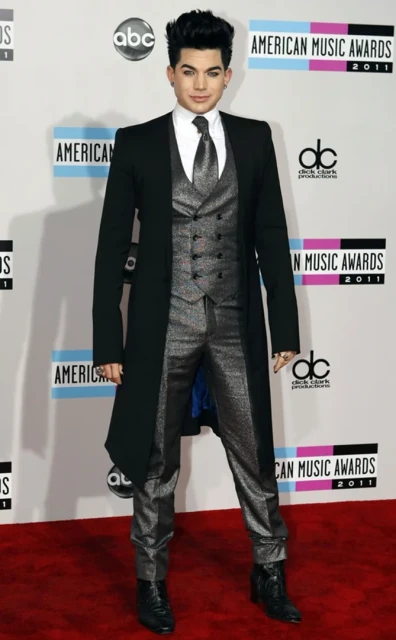 Adam Lambert poses in a glittery grey suit and black overcoat on a red carpet, in front of an American Music Awards backdrop.