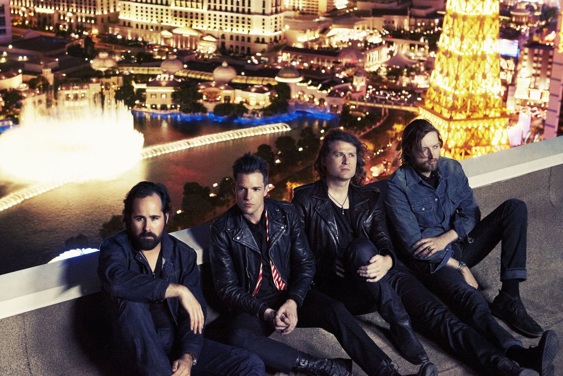 Four adult males sit on a rooftop overlooking a cityscape with bright lights at night, wearing casual attire and leather jackets.
