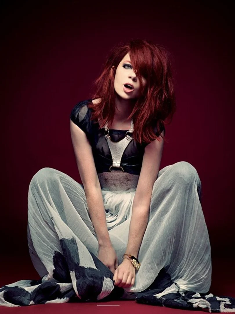 Seated woman with auburn hair poses in black top and light patterned pants against a red background in a fashion photography style.