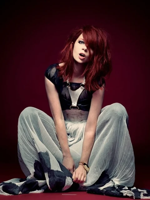 Seated woman with auburn hair poses in black top and light patterned pants against a red background in a fashion photography style.