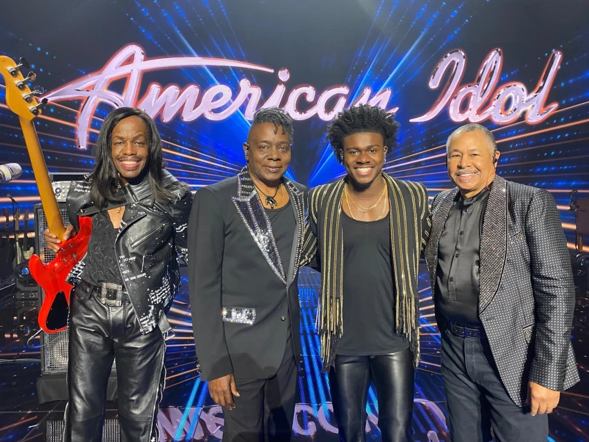 Four musicians posing in a photo studio with stage lighting in front of an American Idol sign. The group is smiling, and one holds a guitar.