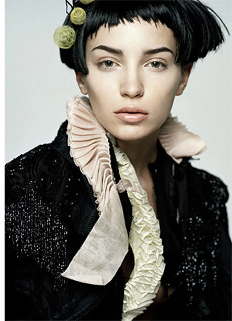 Close-up portrait of a woman with black hair and pale skin wearing a black jacket with ruffles against a light gray background.