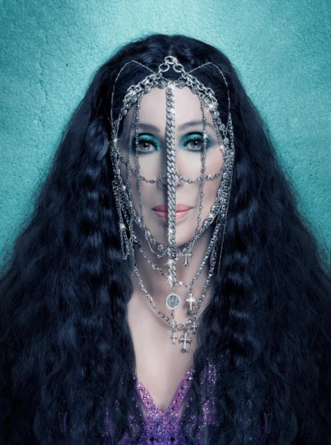 A portrait of Cher wearing a jeweled headpiece and purple sequin top against a textured teal background in a studio setting.
