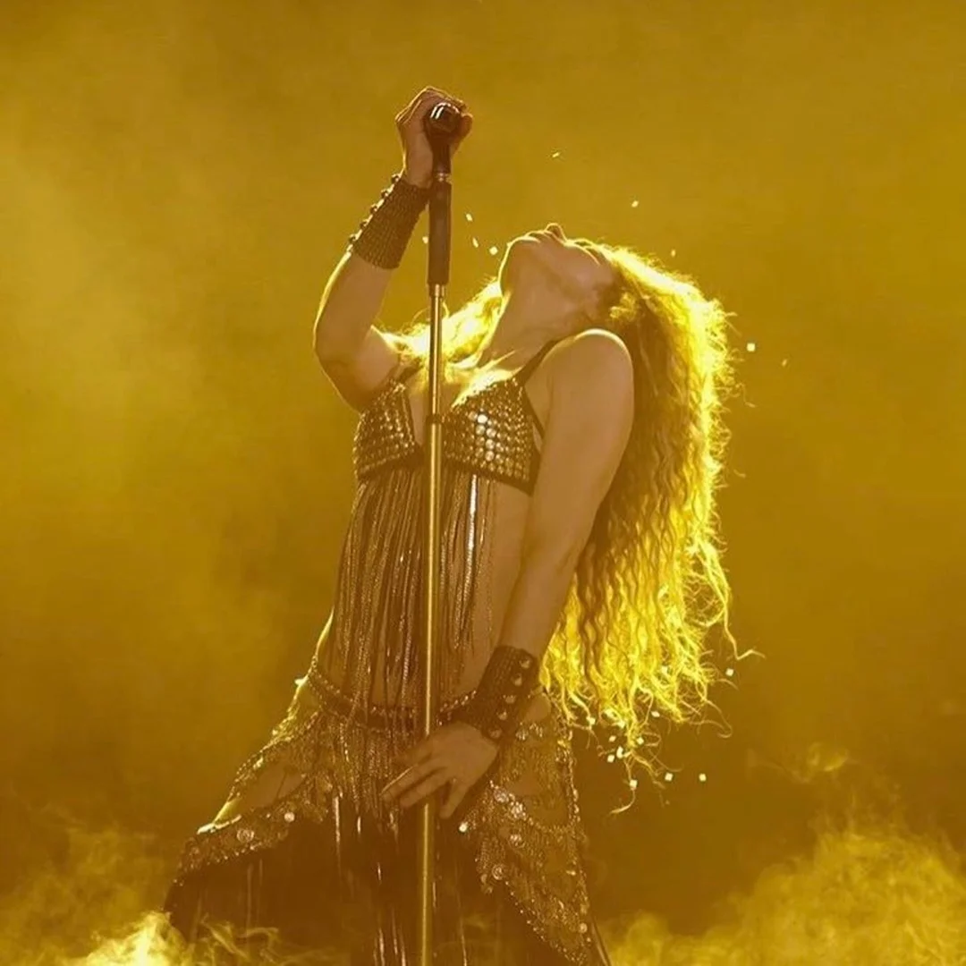 Singer performing on stage holding a microphone, wearing a metallic bra top and skirt with a bright yellow spotlight background.