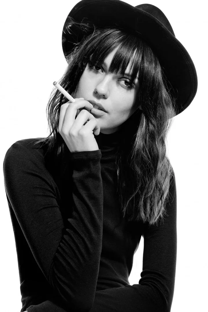 Monochrome portrait of a stylish adult female with a cigarette, wearing a turtleneck sweater and hat against a bright background.